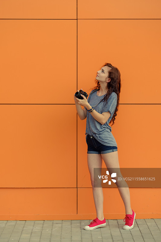 全身肖像，一位美丽的女孩手持老式胶卷相机，仰望着橙色墙壁背景。图片素材