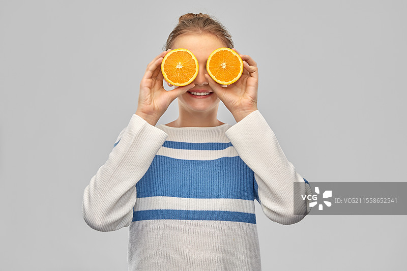 健康饮食、乐趣与人们的概念 - 穿着毛衣的搞笑微笑少女，眼睛是橙子，背景为灰色。微笑的少女眼睛是橙子。图片素材
