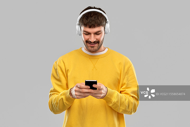 音乐、科技与人们的概念 - 微笑的年轻男子佩戴耳机，手持智能手机，背景为灰色。微笑的年轻男子佩戴耳机，手持智能手机。图片素材