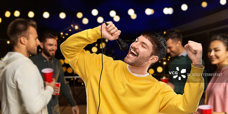 音乐与人们的概念 - 穿着黄色卫衣的年轻男子在夜间屋顶派对背景下用麦克风唱卡拉OK。男子在夜间屋顶派对上用麦克风唱歌。图片素材