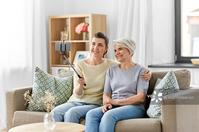 家庭、代际和人们的概念 - 快乐微笑的老年母亲与成年女儿在家中使用自拍杆用智能手机拍照。老年母亲与女儿在家中自拍。图片素材