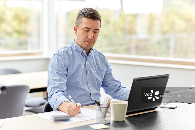 远程工作，商业与人际关系概念 - 中年男子在家办公室使用计算器、文件和笔记本电脑工作。男子在家中使用计算器和文件工作。图片素材