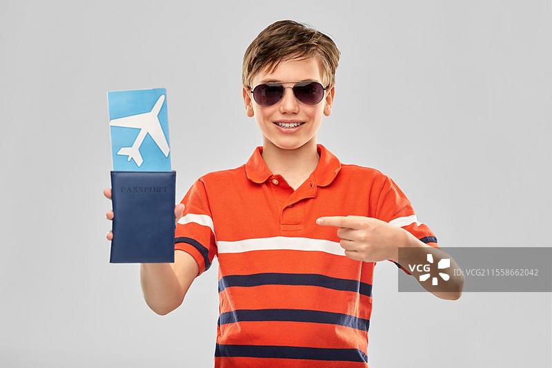 童年、时尚和人们的概念 - 一位快乐微笑的男孩穿着太阳镜和红色 polo 衫，手持机票和护照，背景为灰色。戴着太阳镜的快乐男孩手持机票和护照。图片素材