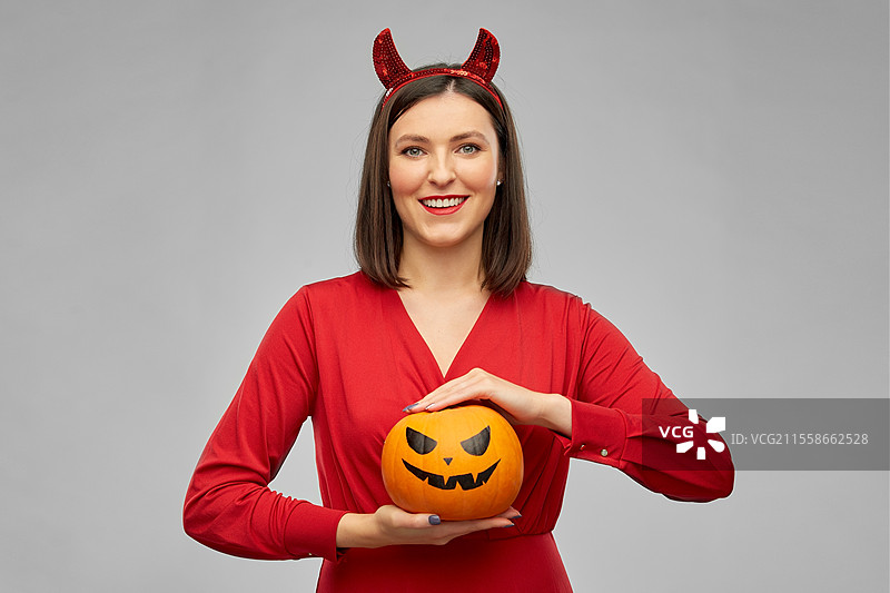 节日、主题派对和人们的概念 - 开心微笑的女人穿着红色的万圣节恶魔服装，头戴角，手持南瓜灯，背景为灰色。穿着恶魔服装的女人与南瓜灯。图片素材