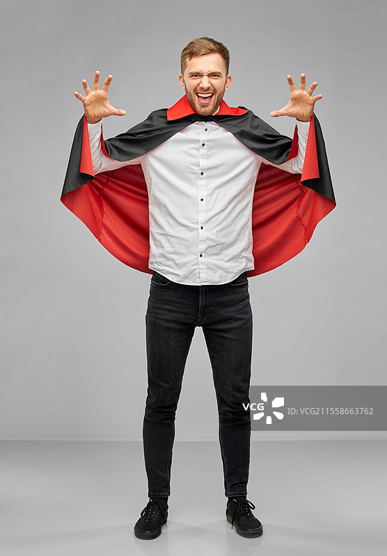 假期、主题派对和人们的概念 - 穿着吸血鬼和德古拉斗篷的男人在灰色背景上吓人。穿着吸血鬼服装的男人在吓人。图片素材