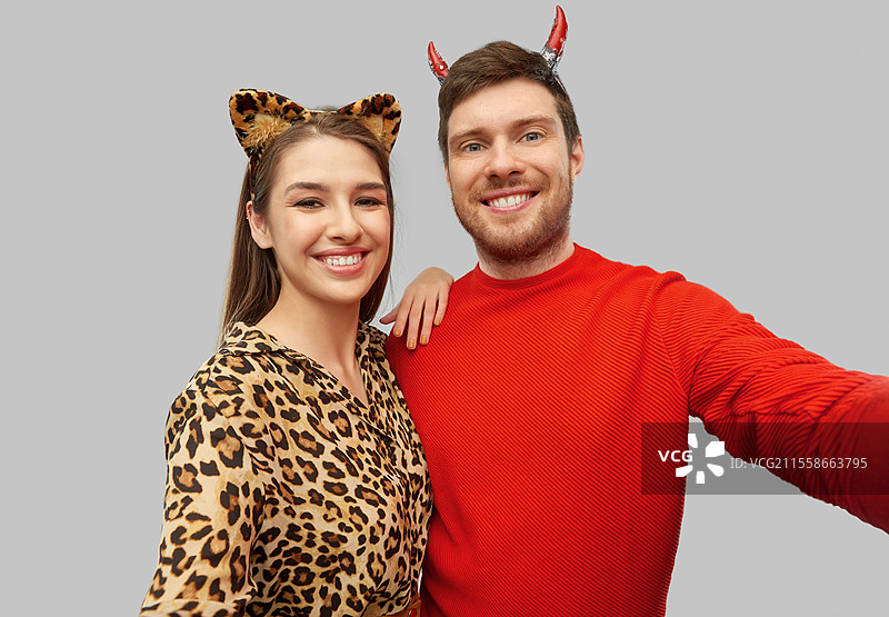 假期和人们的概念 - 一对快乐微笑的情侣穿着恶魔和豹子的万圣节服装在灰色背景下自拍。快乐的情侣在万圣节服装中自拍。图片素材