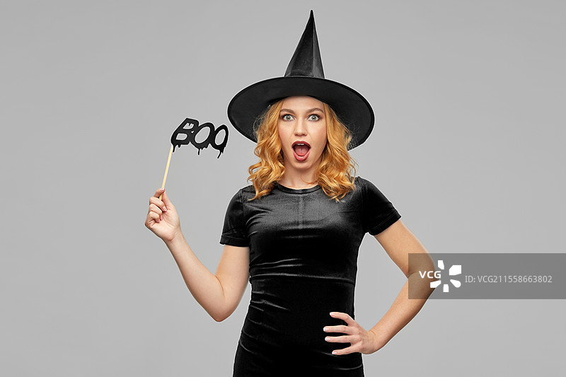 假日和照片亭概念 - 穿着黑色万圣节女巫服装、手持派对道具的快乐微笑女性，背景为灰色。穿着女巫服装的女性与道具。图片素材