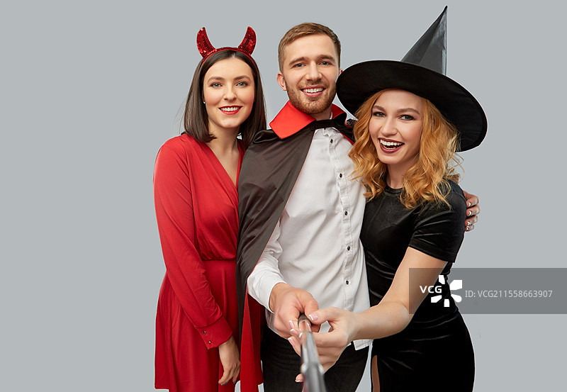 友谊、假期和人们的概念 - 一群快乐微笑的朋友穿着女巫、恶魔和吸血鬼的万圣节服装，在灰色背景下用自拍杆拍照。快乐的朋友们在万圣节服装中自拍。图片素材