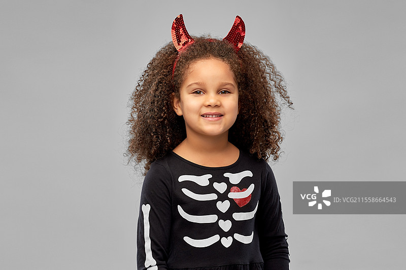 万圣节，节日和童年概念 - 微笑的非裔美国女孩穿着黑色服装和红色魔鬼角，背景为灰色。女孩在万圣节穿着黑色裙子和魔鬼角。图片素材