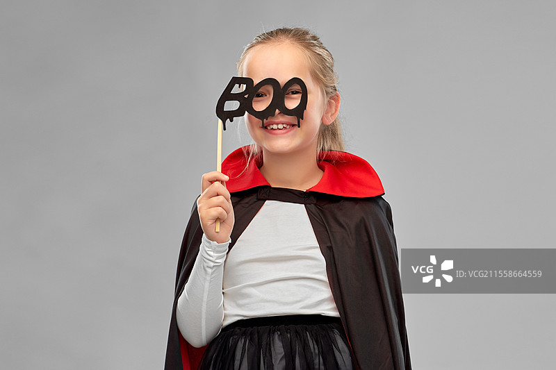 万圣节，节日和童年概念 - 穿着黑色斗篷的德古拉装扮女孩在灰色背景下手持派对道具。万圣节时穿着德古拉服装的女孩。图片素材