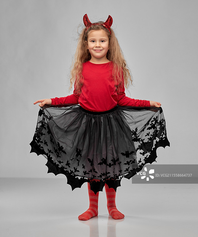 万圣节，节日和童年概念 - 微笑的女孩穿着派对服装，头戴红色魔鬼角，背景为灰色。女孩穿着万圣节服装和魔鬼角。图片素材