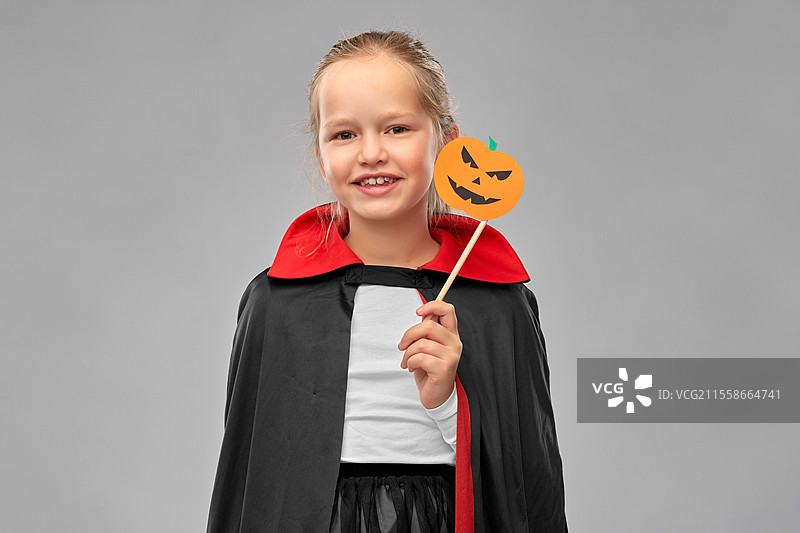 万圣节、节日和童年概念 - 穿着黑色斗篷的德古拉装扮女孩，手持南瓜灯派对配饰，背景为灰色。万圣节时穿着德古拉服装的女孩。图片素材