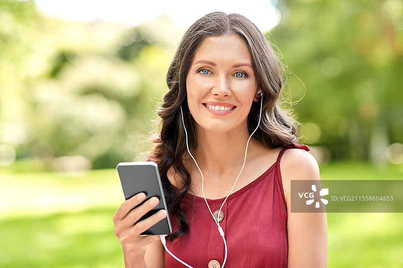 休闲与人们的概念 - 在夏季公园中，快乐微笑的女性手持智能手机，佩戴耳机，聆听音乐。公园中的女性与智能手机和耳机。图片素材
