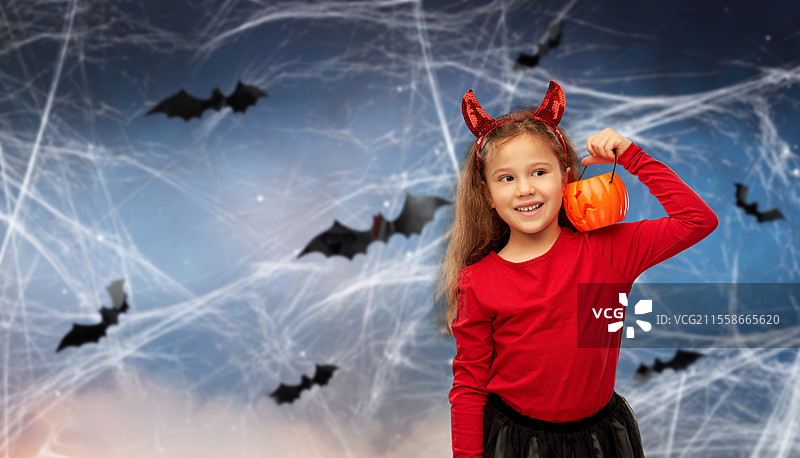 万圣节、节日和童年概念 - 微笑的女孩穿着派对服装，头戴红色魔鬼角，手持南瓜灯，背景是夜空中的蝙蝠和蜘蛛网。女孩穿着万圣节服装，手拿南瓜灯。图片素材