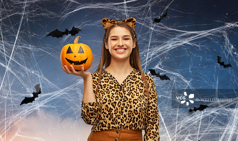 假期、主题派对和人们的概念 - 开心微笑的女性穿着豹纹万圣节服装，头戴耳朵，手持南瓜灯，背景是星空中飞舞的蜘蛛网和蝙蝠。穿着豹纹万圣节服装的女性与南瓜灯。图片素材