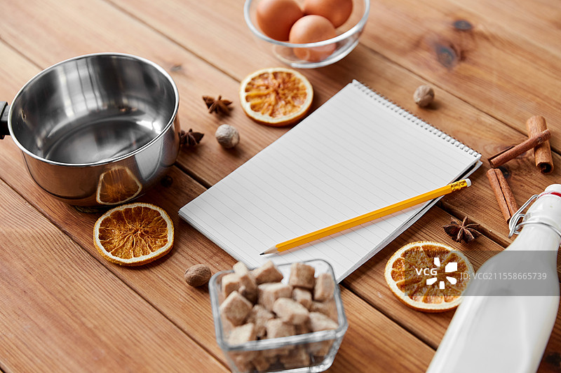 烹饪、圣诞节和季节性饮品概念 - 木质背景上的笔记本与铅笔、蛋奶酒的配料、锅。笔记本、铅笔、蛋奶酒烹饪的配料。图片素材