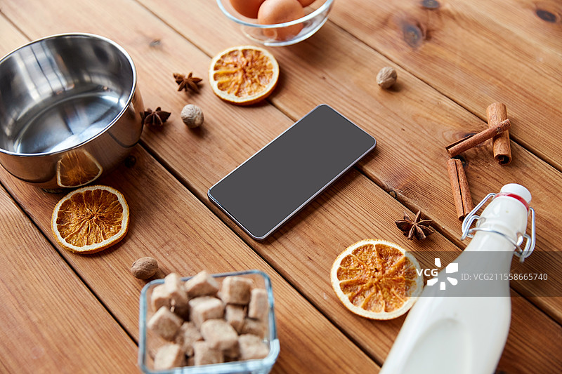 烹饪、圣诞节和季节性饮品概念 - 智能手机、蛋奶酒的配料、木质背景上的锅。智能手机、蛋奶酒烹饪的配料。图片素材
