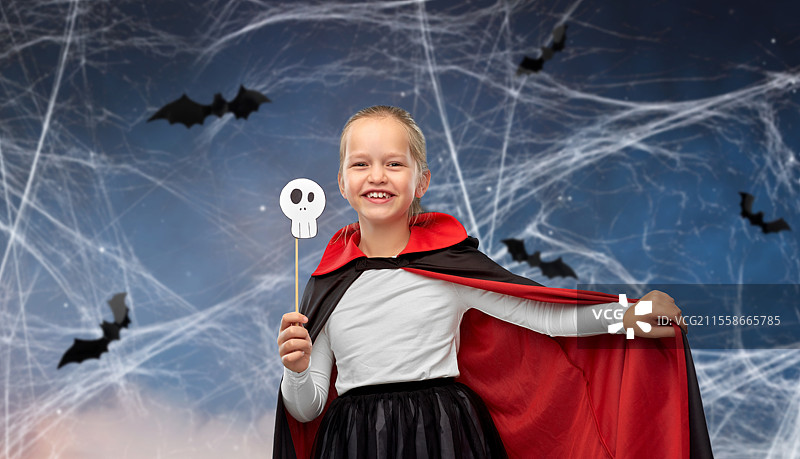 万圣节、节日和童年概念 - 穿着黑色斗篷的德古拉装扮女孩在夜空中手持骷髅派对道具，背景有蝙蝠和蛛网。万圣节时穿着德古拉服装的女孩。图片素材