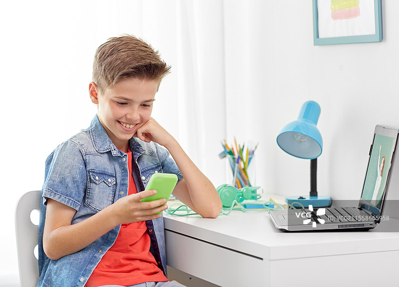 教育、电子学习和学校概念 - 开心微笑的男孩手持智能手机，老师在笔记本电脑上进行在线课程，家中书桌前。开心的男孩在家中使用智能手机和笔记本电脑。图片素材