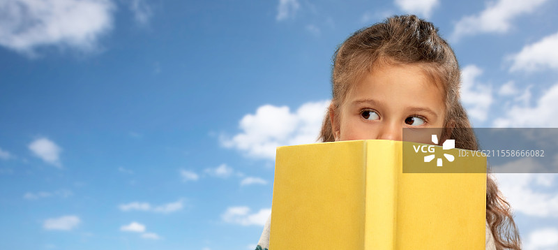 阅读、教育和童年概念 - 小女孩躲在黄色书本后面，背景是蓝天和白云。小女孩躲在书本后面，背景是天空和云朵。图片素材