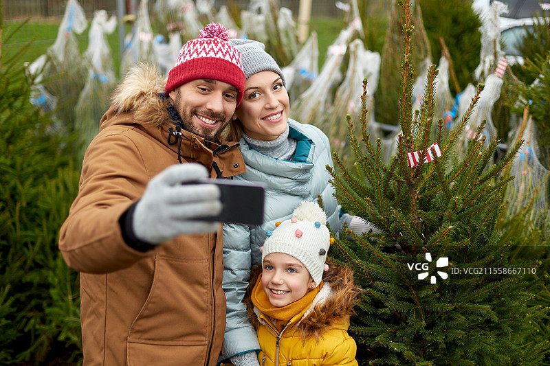 家庭、冬季假期和人们的概念 - 快乐的母亲、父亲和小女儿在街市上购买圣诞树并用智能手机自拍。家庭在市场上与圣诞树合影。图片素材