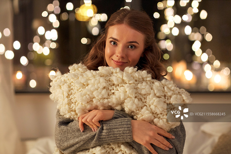 圣诞节、假日和人们的概念 - 快乐的年轻女性在家中卧室的床上，夜晚背景中有节日灯光，手中抱着柔软的枕头。图片素材