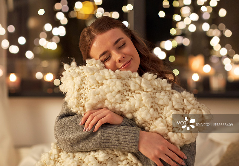 圣诞节、假日和人们的概念 - 快乐的年轻女性在家中卧室的床上，夜晚背景中有节日灯光，手中抱着柔软的枕头。图片素材