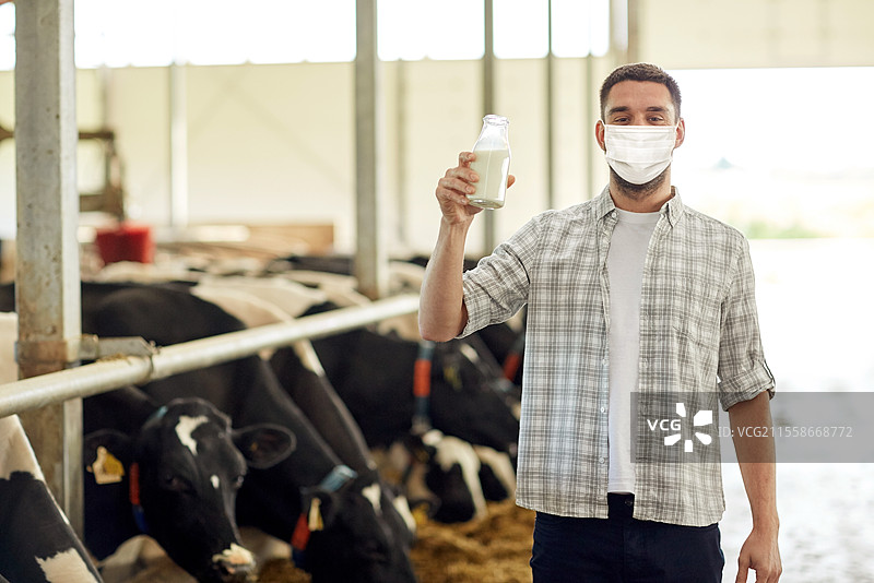 农业行业，农业，人与动物饲养概念 - 年轻男子或农民在奶牛舍里拿着一瓶牛奶，佩戴医用口罩以防病毒疾病。在奶牛场的男子或农民戴着口罩，手持牛奶。图片素材