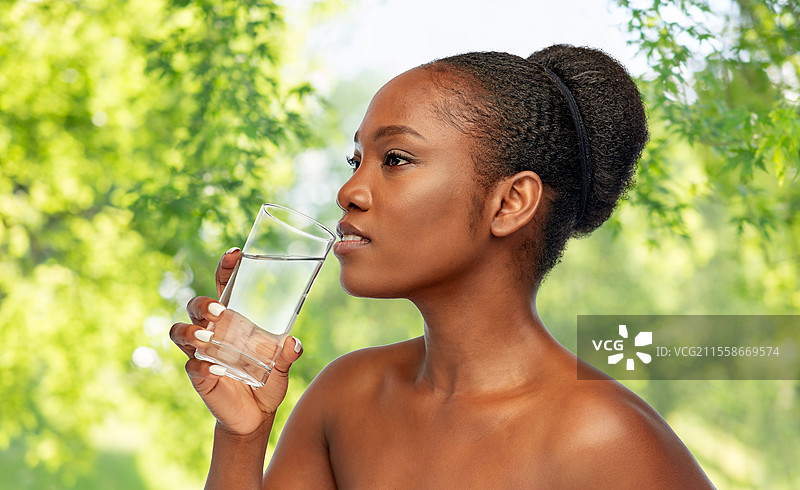 美丽与人文概念 - 年轻非洲裔美国女性的肖像，肩膀，手持玻璃杯饮水，背景为绿色自然景观。年轻非洲裔美国女性与水杯。图片素材