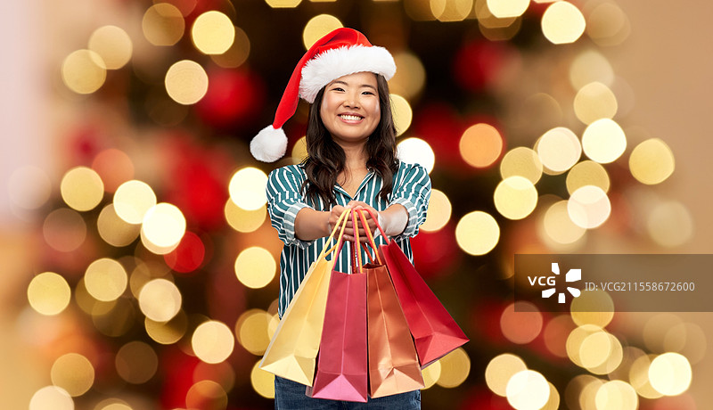 冬季假期和促销概念 - 开心的亚洲年轻女性戴着圣诞助手的帽子，手提购物袋，背景是圣诞树。开心的亚洲女性手提购物袋庆祝圣诞节。图片素材
