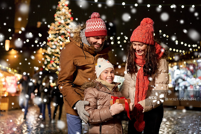 家庭、冬季假期和庆祝概念 - 快乐的母亲、父亲和小女儿在爱沙尼亚塔林市政厅广场的圣诞市场上，雪中与礼物同在。快乐的家庭在城市的圣诞市场上与礼物同在。图片素材