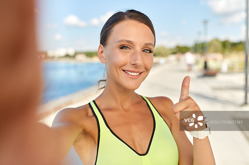 人们、运动和健身概念 - 微笑的年轻运动女性在海滨长廊户外自拍并竖起大拇指。运动女性自拍并竖起大拇指。图片素材