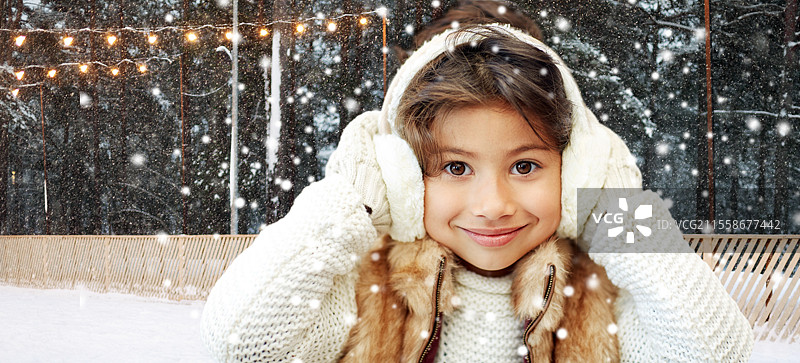 圣诞节、季节和儿童概念 - 快乐的小女孩戴着耳罩，背景是冬季滑冰场。冬季滑冰场上的女孩戴着耳罩。图片素材