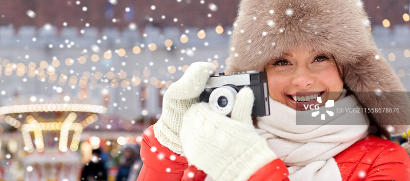人们、科技和假日概念 - 戴着冬季毛皮帽的快乐女性在圣诞市场背景下用胶卷相机拍照。快乐女性在圣诞市场前拿着胶卷相机。图片素材