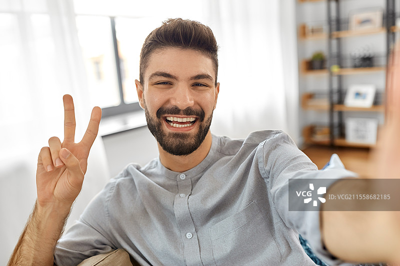科技、人与生活方式的概念 - 快乐的男人在家中自拍或进行视频通话，并做出和平手势。男人在家中自拍或进行视频通话。图片素材