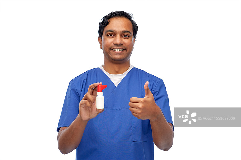 医学、职业和医疗保健概念 - 穿着蓝色制服的快乐微笑的印度医生或男护士，手持口腔喷雾，竖起大拇指，背景为白色。微笑的印度男性医生或护士与药物。图片素材