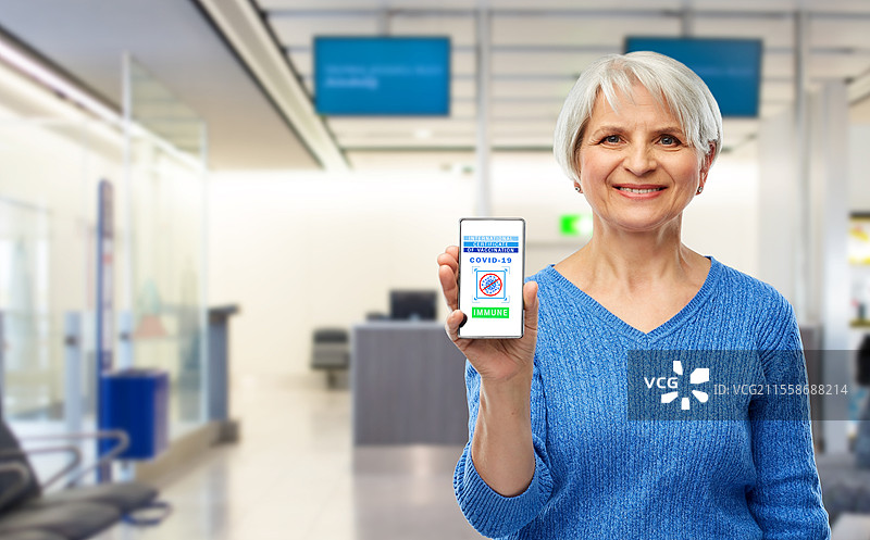 安全旅行、科技与医疗保健概念 - 开心微笑的老年女性在机场背景下手持并展示手机，手机屏幕上显示国际疫苗接种证明。老年女性手持手机上的疫苗接种证明。图片素材
