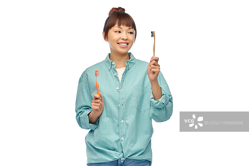 牙齿护理、口腔卫生和人们的概念 - 一位穿着青绿色衬衫的年轻亚洲女性在白色背景前微笑着的肖像，正在选择木制和塑料牙刷。手中拿着木制和塑料牙刷的亚洲女性。图片素材