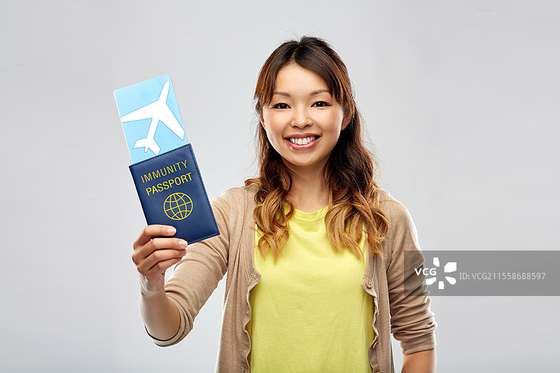 安全旅行、旅游和医疗保健概念 - 开心微笑的年轻亚洲女性手持机票和免疫护照，背景为灰色。亚洲女性手持机票和免疫护照。图片素材