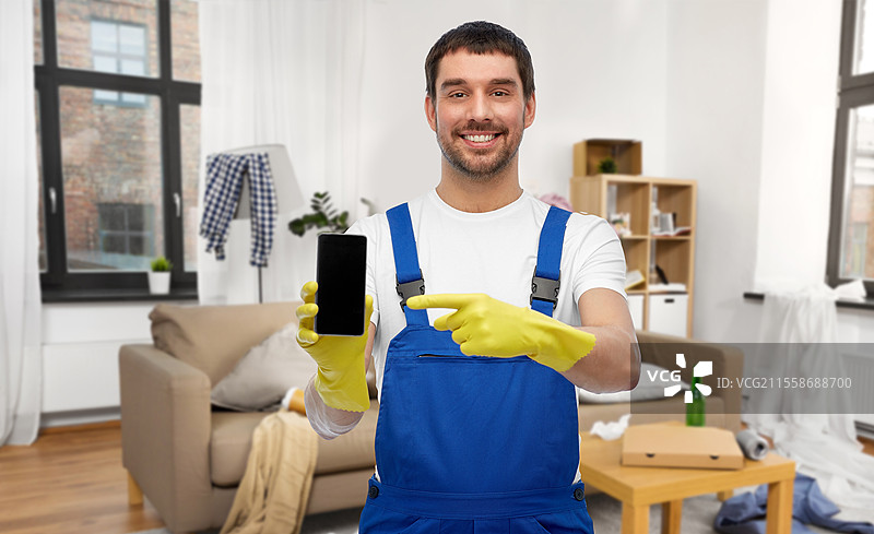 职业、清洁服务和人们的概念 - 开心微笑的男性工人或清洁工穿着工作服和手套，在家居背景下展示智能手机。男性工人或清洁工在家中展示智能手机。图片素材