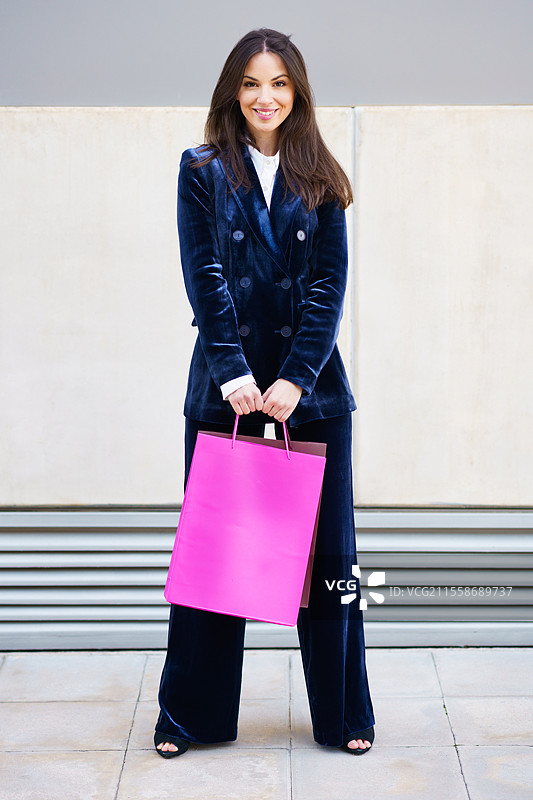 年轻女性穿着蓝色西装，手提多个购物袋，靠近购物中心。生活方式概念。图片素材