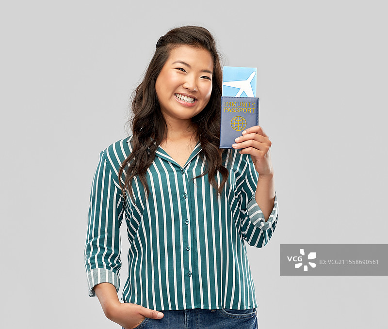 安全旅行、旅游和医疗保健概念 - 开心微笑的年轻亚洲女性手持机票和免疫护照，背景为灰色。亚洲女性手持机票和免疫护照。图片素材