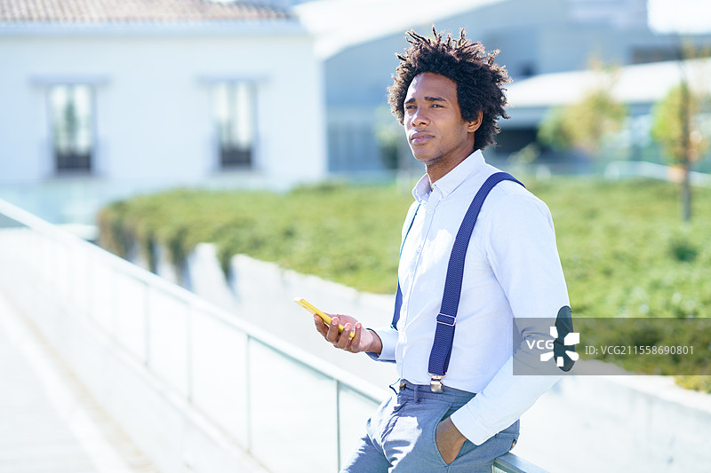 一位留着非洲卷发的黑人男子在办公楼附近使用智能手机。穿着衬衫和吊带裤的卷发男士。图片素材