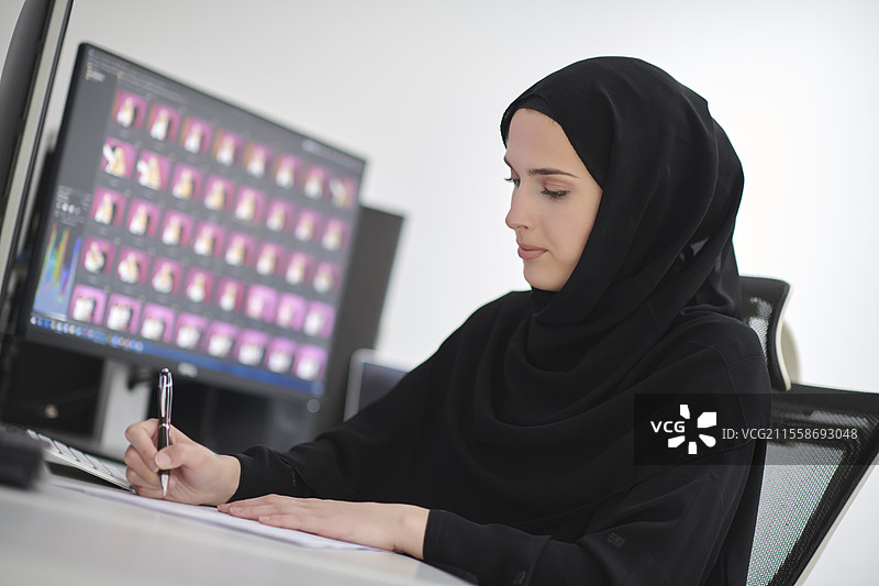 身穿头巾的穆斯林女性平面设计师在办公室使用图形平板和两台显示器进行电脑工作，正在编辑照片。图片素材
