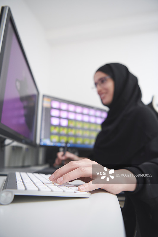 身穿头巾的穆斯林女性平面设计师在办公室使用图形平板和两台显示器进行电脑工作，正在编辑照片。图片素材