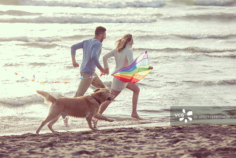 年轻情侣在秋天的海滩上玩耍，和狗狗一起放风筝。图片素材