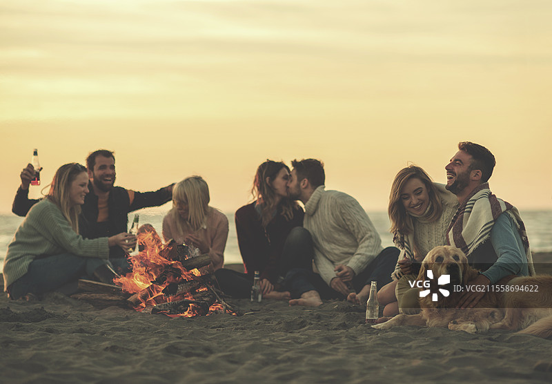 快乐无忧的年轻朋友们在海滩的篝火旁享受乐趣，喝着啤酒，夕阳开始西沉。图片素材
