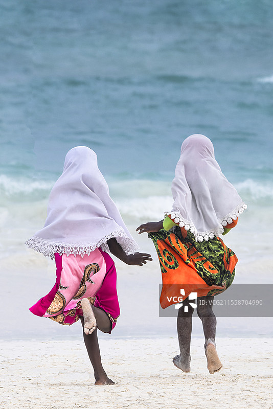 两个小女孩穿着色彩斑斓的裙子和白色头巾，朝大海奔跑。在傍晚时分，来自坦桑尼亚桑给巴尔东海岸一个小村庄的孩子们来到白色沙滩和碧蓝海水上玩耍。图片素材