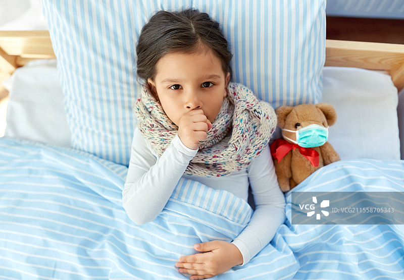 健康、儿童和疫情概念 - 生病咳嗽的女孩躺在床上，抱着戴着防护口罩的泰迪熊玩具。生病咳嗽的女孩与泰迪熊躺在床上。图片素材