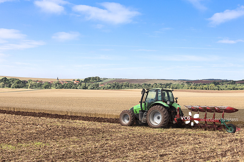 在法国洛林，远处的村庄中，教堂被红瓦房屋环绕，正在用拖拉机和可逆五铧犁耕作小麦地，经过秸秆耕作。图片素材